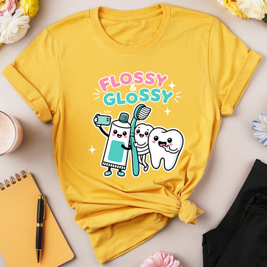 A yellow graphic t-shirt with a cute cartoon toothbrush and tooth design that reads 'FLOSSY GLOSSY' in pink and blue. The t-shirt is styled in a flat lay with black pants, a yellow notebook, a pink drink, and flowers on a light beige background.