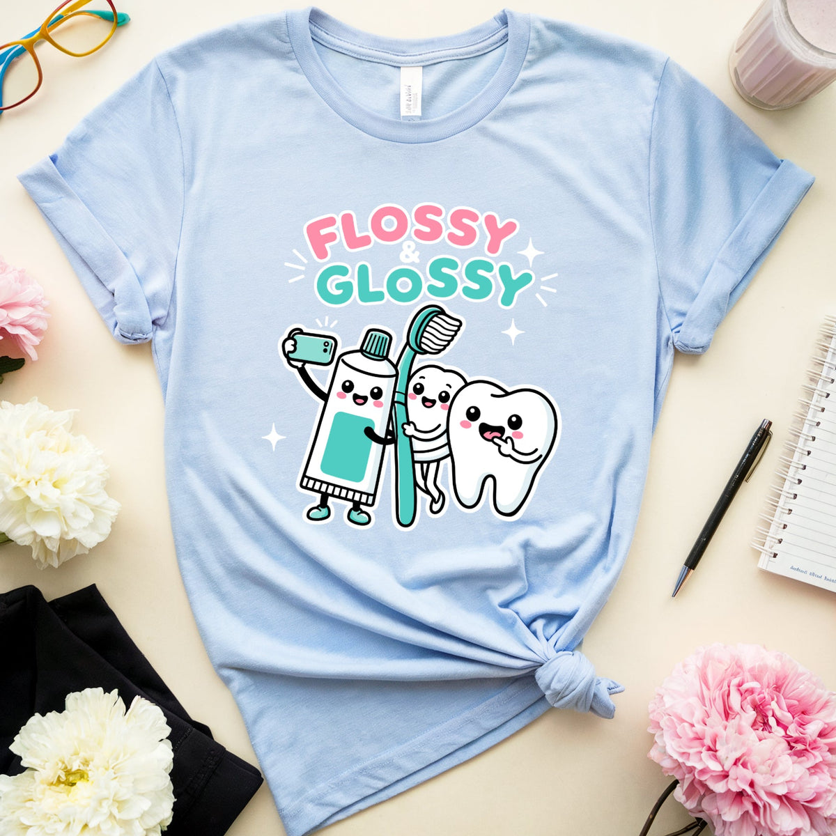 A light blue graphic t-shirt with a cartoon design of a toothbrush, tooth, and smiling tooth surrounded by the words 'FLOSSY GLOSSY'. The t-shirt is styled in a flat-lay arrangement on a beige background with pink and white flowers, a pair of glasses, a notebook, a pen, and a drink.