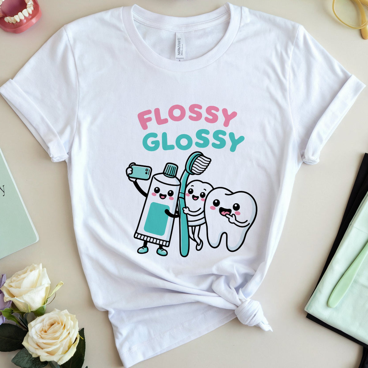 A white short-sleeve graphic T-shirt laid flat on a light beige surface, styled with flowers, glasses, a model mouth, a notebook, and a folded toothbrush case. The shirt has a colorful cartoon graphic reading “FLOSSY GLOSSY” with a toothpaste tube, toothbrush, and tooth character, plus a tied hem.