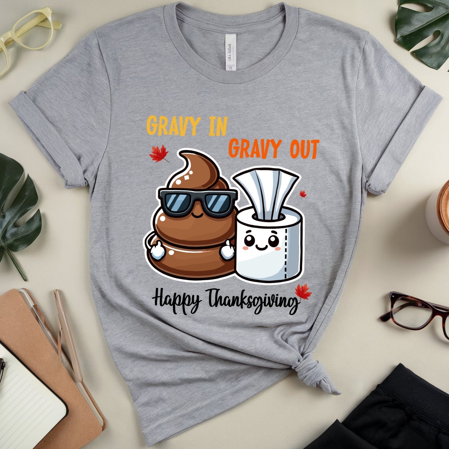 Gray t-shirt with cartoon design of gravy and paper towels, text 'Gravy in Gravy Out Happy Thanksgiving', on a light surface with accessories.
