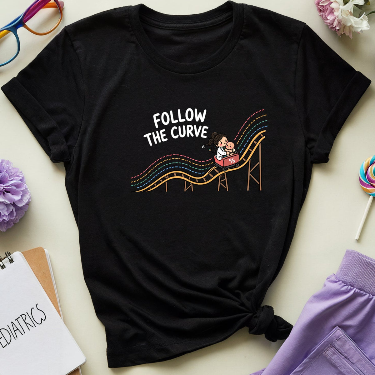 A black graphic T-shirt laid flat on a light beige surface, surrounded by styled props including purple flowers, rainbow glasses, a spiral notebook, a lollipop, and lavender pants. The shirt has a colorful cartoon slide graphic with the text 'FOLLOW THE CURVE'.