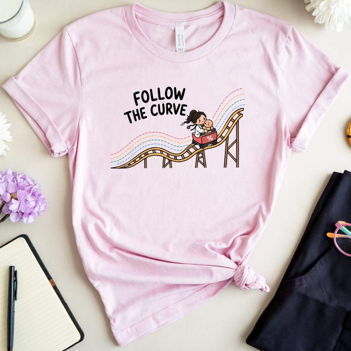 A light pink graphic t-shirt featuring the text 'FOLLOW THE CURVE' and a cartoon roller coaster illustration, styled in a flat lay with a notebook, glasses, a cookie, and flowers on a beige background.