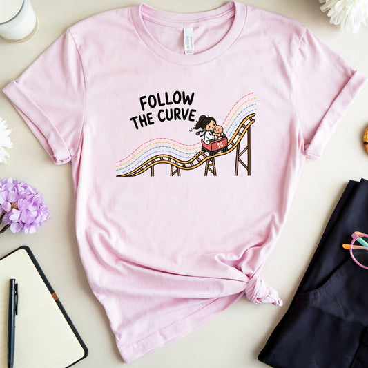 A light pink graphic t-shirt featuring the text 'FOLLOW THE CURVE' and a cartoon roller coaster illustration, styled in a flat lay with a notebook, glasses, a cookie, and flowers on a beige background.
