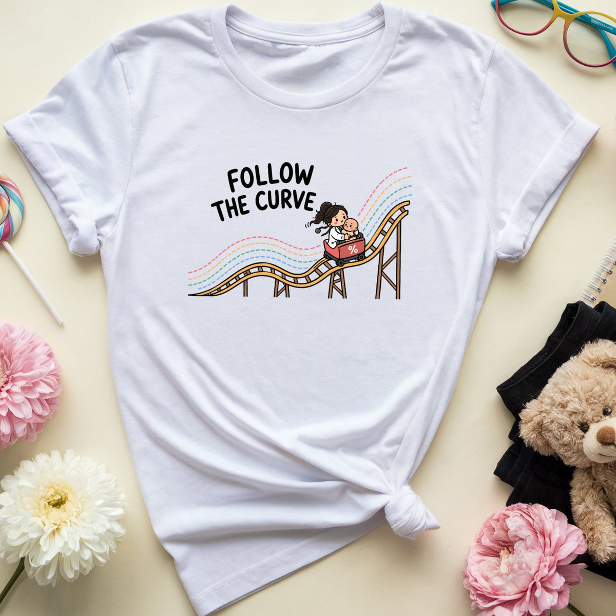 A white graphic t-shirt with a cartoon roller coaster design and the text 'FOLLOW THE CURVE' is styled in a flat lay with pink and white flowers, a teddy bear, glasses, and a notebook on a light beige background.