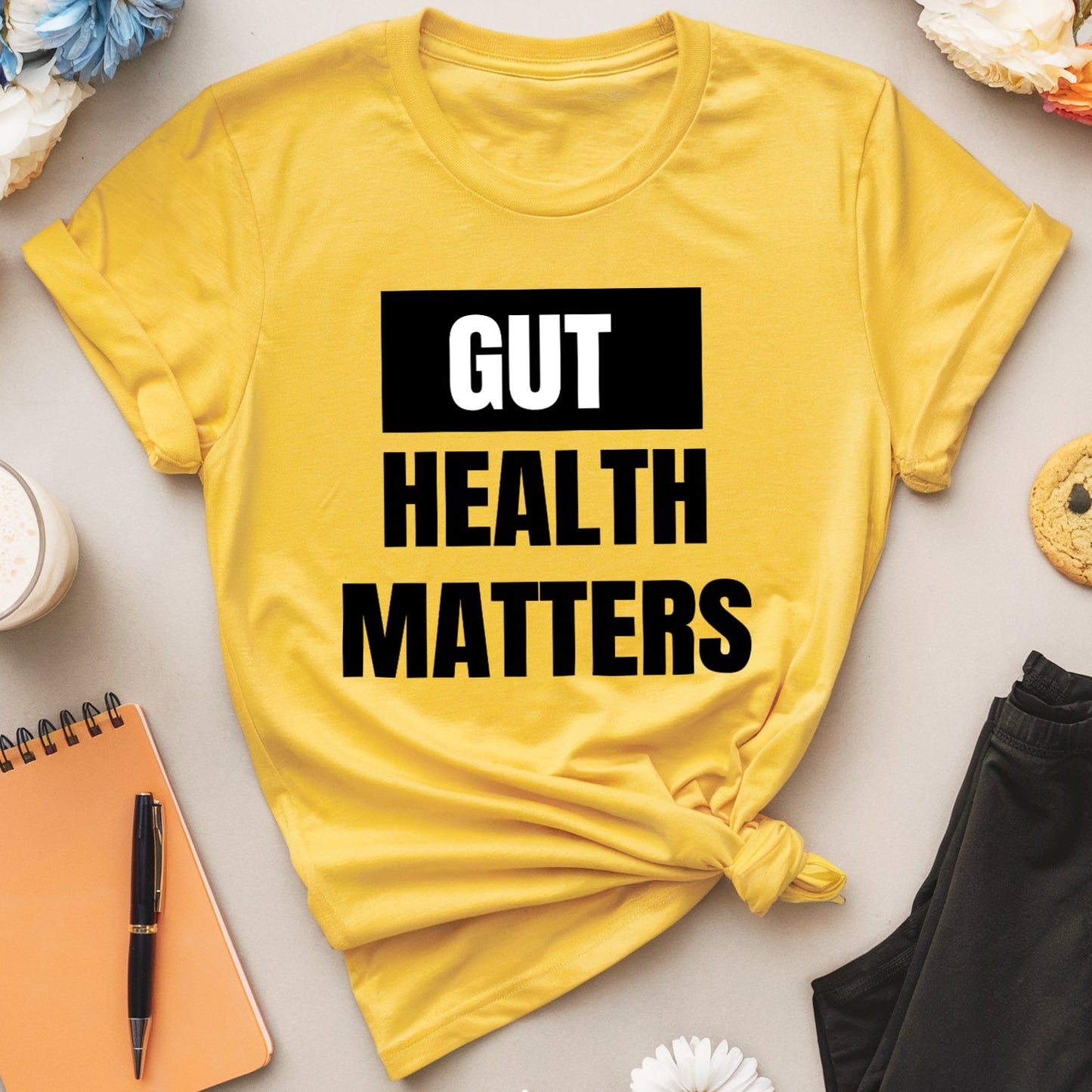 Yellow t-shirt with 'GUT HEALTH MATTERS' text on a table with flowers, cookies, and a notebook.