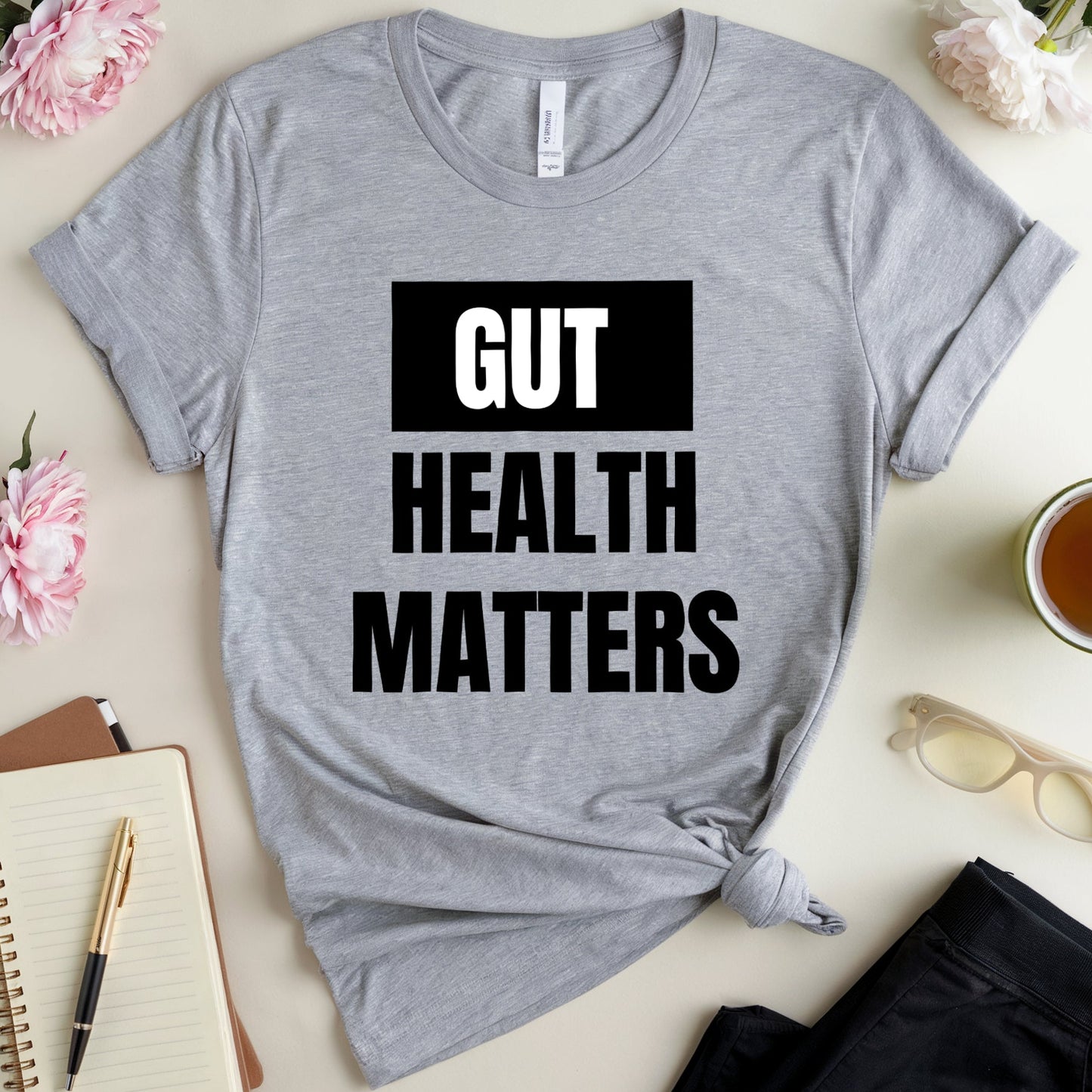 Gray t-shirt with 'GUT HEALTH MATTERS' text on a table with flowers, notebook, and tea.