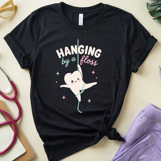 A black short-sleeve graphic T-shirt laid flat on a light beige surface, styled with dental-themed props including a stethoscope, glasses, a pen, a notebook, and folded purple scrubs. The shirt has a cartoon tooth character hanging from a floss handle with the text 'HANGING by a floss' in white and pink.
