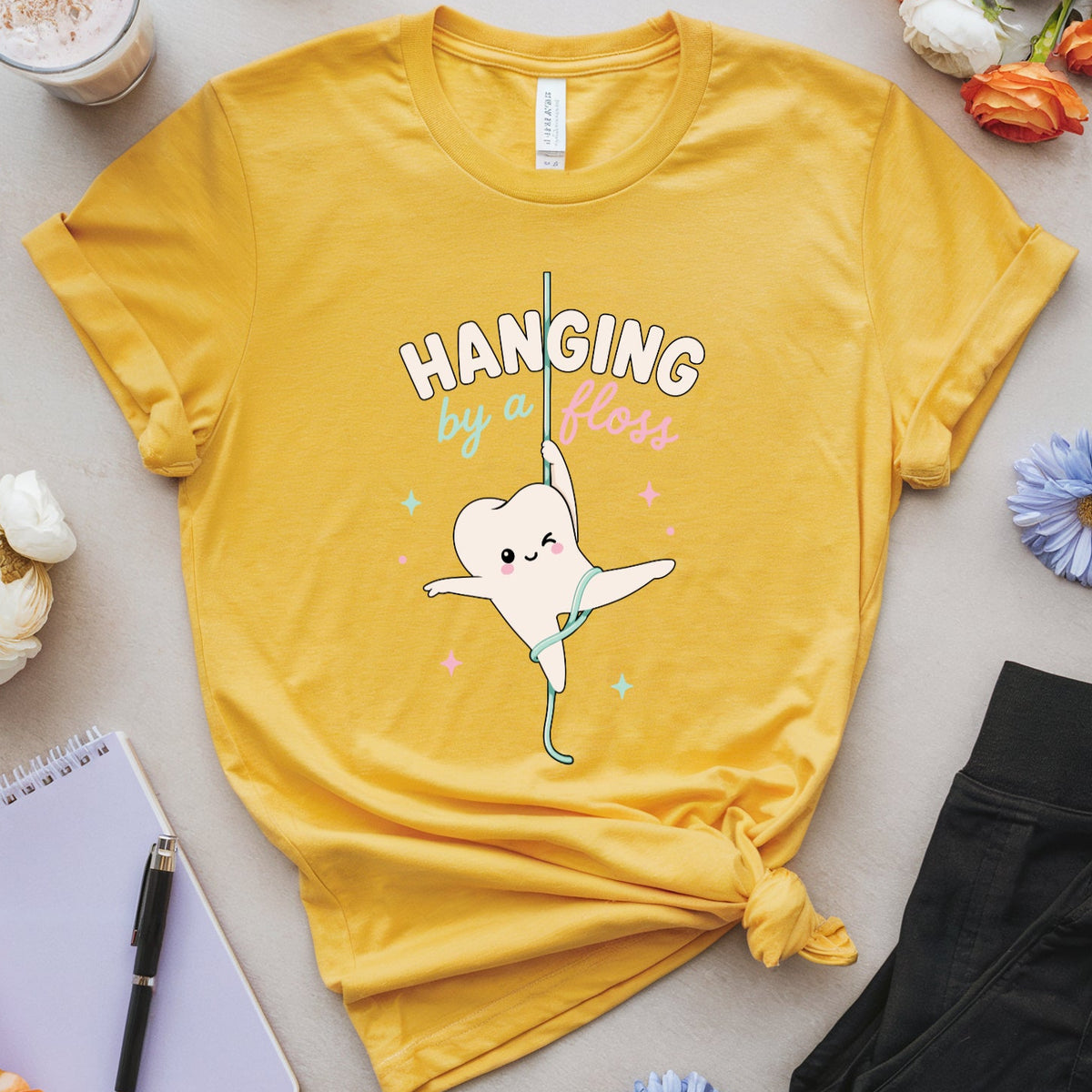 A yellow graphic t-shirt featuring a cartoon tooth character and the text 'HANGING by a floss' in a styled flat-lay arrangement with flowers, glasses, a notebook, and a drink.