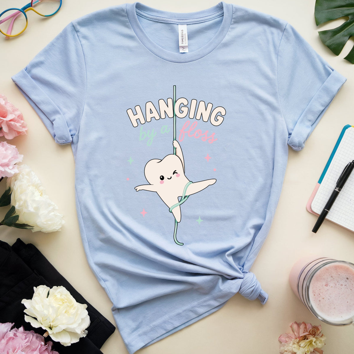 A light blue graphic t-shirt featuring a cartoon tooth character hanging from a bar with the text 'HANGING Tooth' in white and pink. The t-shirt is styled in a flat lay on a beige surface with pink and white flowers, a pair of colorful glasses, a notebook, a black cloth, and a drink.