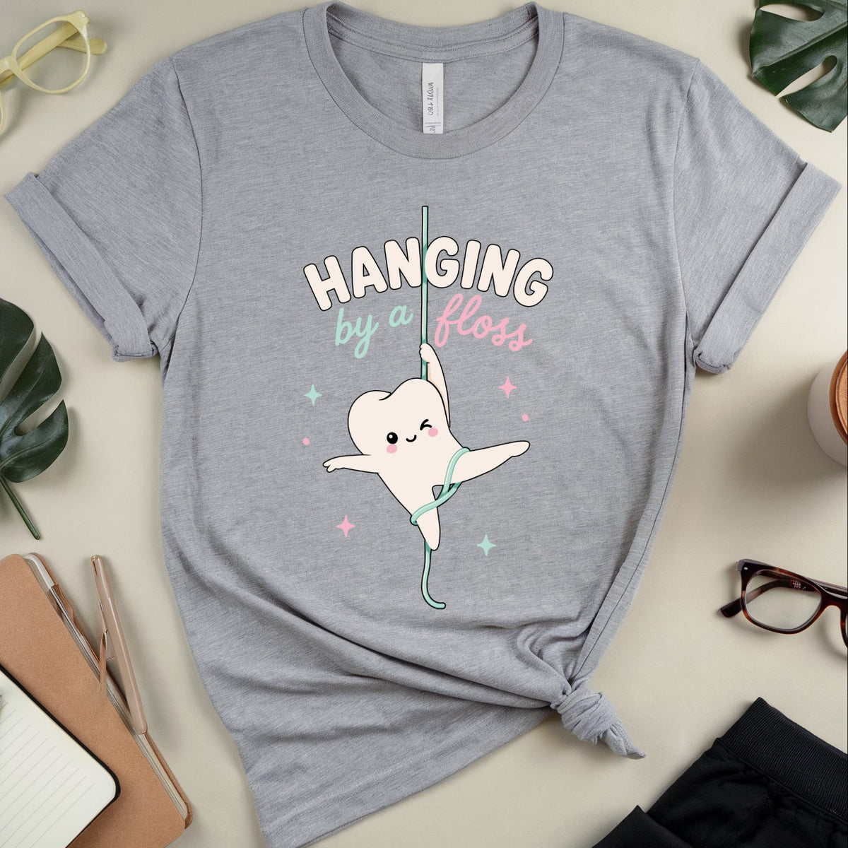 A heather gray graphic t-shirt featuring a cartoon tooth character hanging by a floss, with the text 'HANGING by a Floss' in white and pastel pink and green. The t-shirt is styled in a flat lay with glasses, a notebook, a coffee cup, and green leaves on a light beige background.