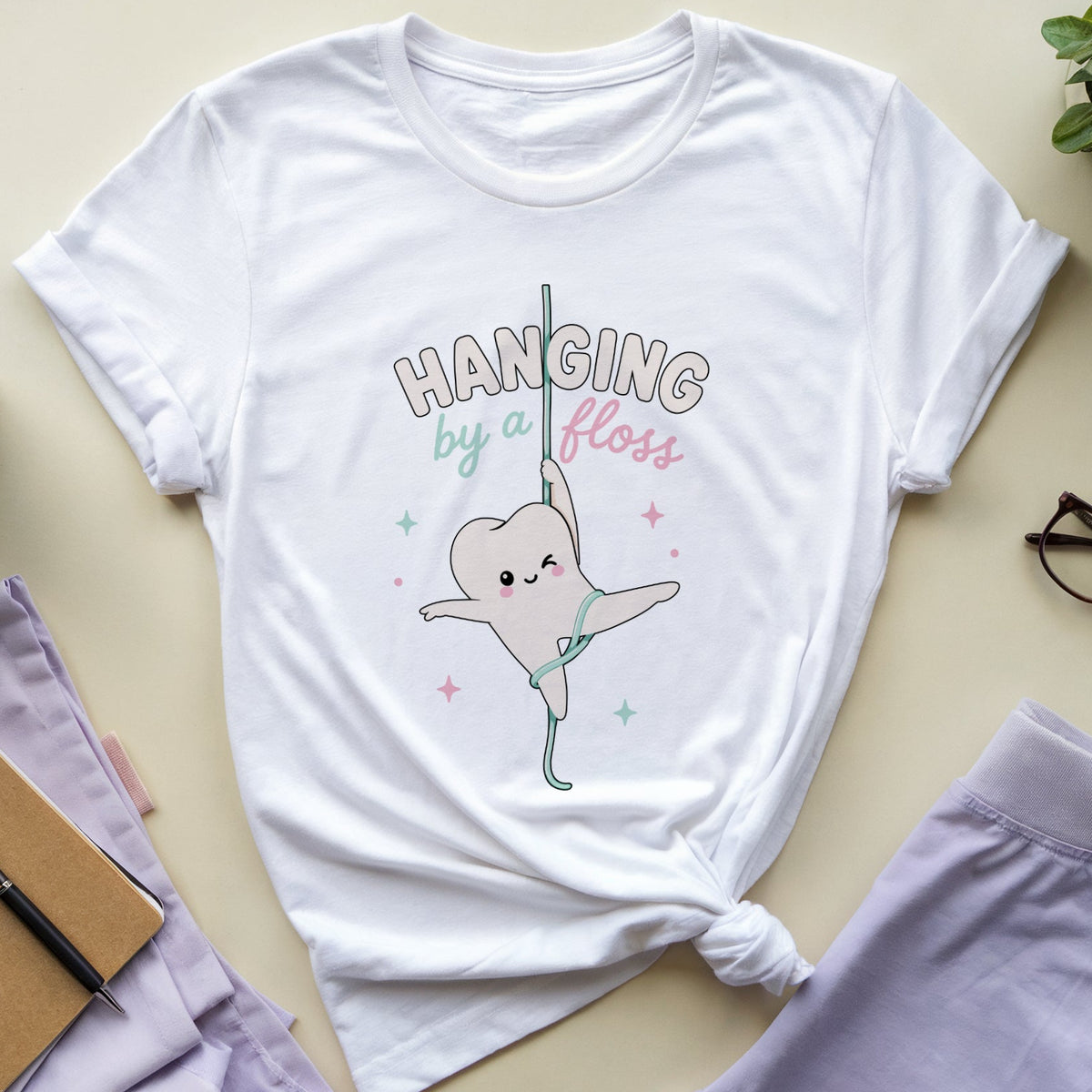 A white graphic t-shirt featuring a cute cartoon tooth gymnast hanging from a bar with the text 'HANGING by a bless'. The t-shirt is styled in a flat-lay arrangement on a beige background with lavender pants, glasses, a notebook, and a plant.
