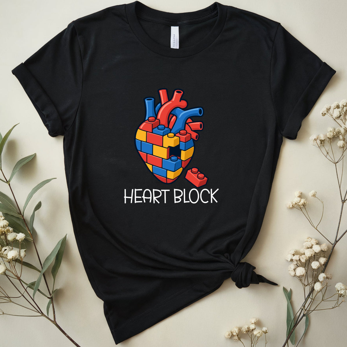 A black short-sleeve graphic T-shirt laid flat on a light beige background with dried flowers around it. The shirt has a colorful heart-shaped block pattern on the chest with the words 'HEART BLOCK' printed below in white.