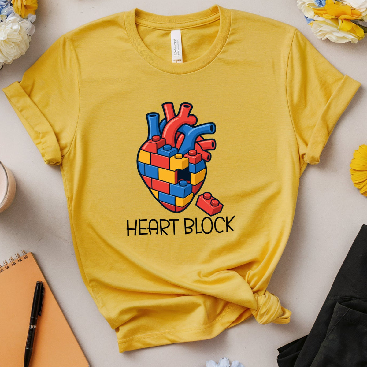 A yellow graphic t-shirt featuring a colorful heart made of red, blue, and yellow building blocks with the text 'HEART BLOCK' underneath. The t-shirt is styled in a flat-lay arrangement with flowers, glasses, a notebook, and a pair of black pants on a light beige background.