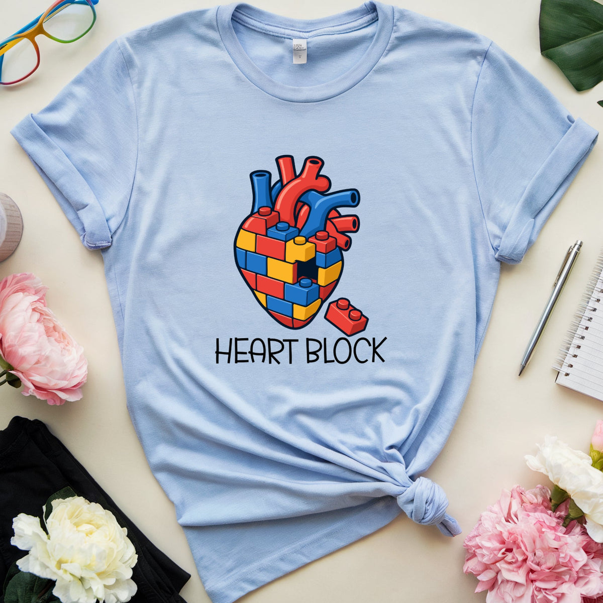 A light blue graphic t-shirt with a colorful heart made of puzzle pieces and the text 'HEART BLOCK' underneath, styled in a flat lay with pink flowers, glasses, a notebook, and a cup on a beige background.