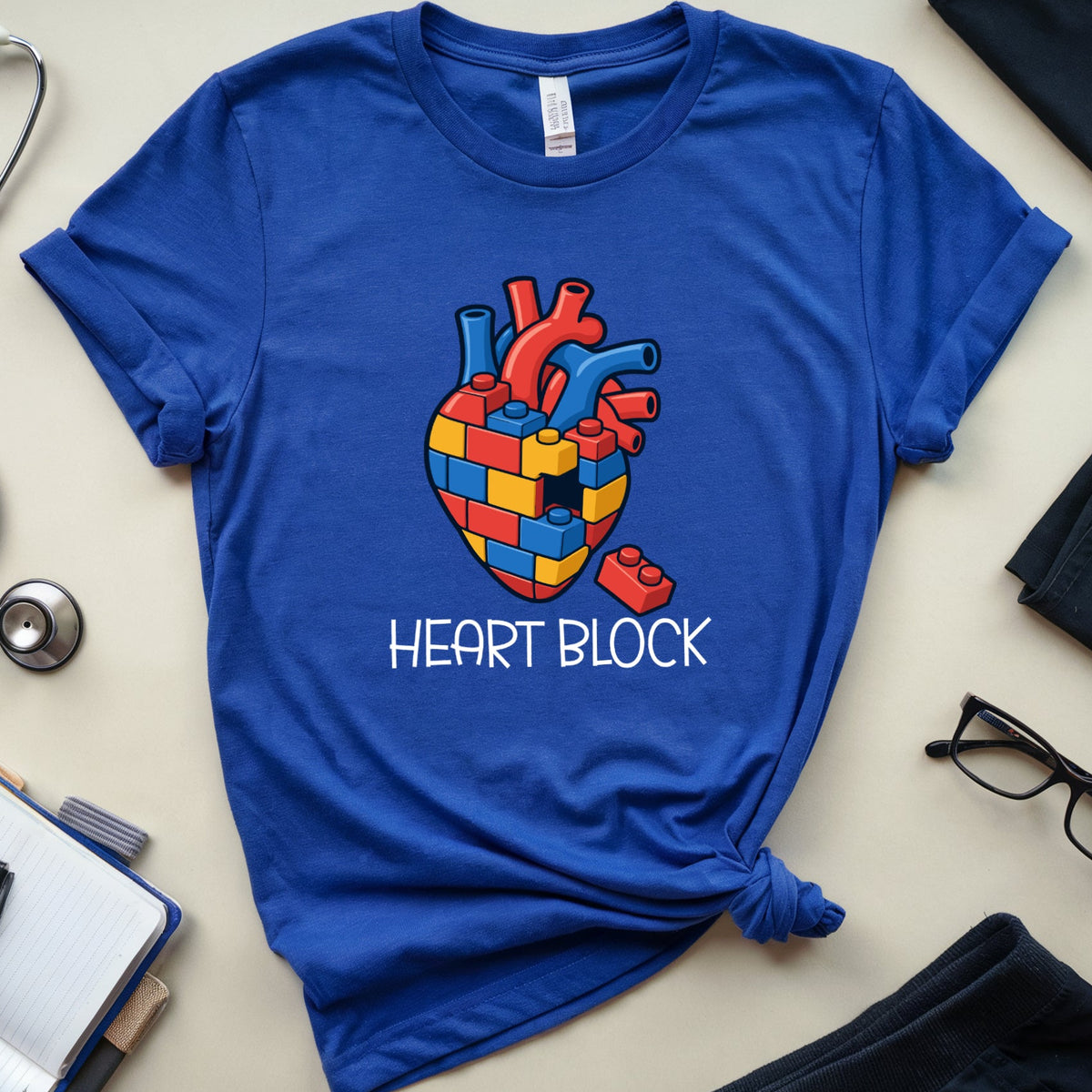 A royal blue short-sleeve t-shirt with a graphic of a colorful heart made of medical blocks and the text 'HEART BLOCK' printed below it. The t-shirt is styled in a flat lay on a light beige surface with a stethoscope, notebook, glasses, and clothing items around it.