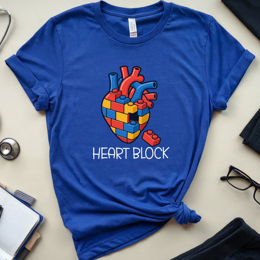 A royal blue short-sleeve t-shirt with a graphic of a colorful heart made of medical blocks and the text 'HEART BLOCK' printed below it. The t-shirt is styled in a flat lay on a light beige surface with a stethoscope, notebook, glasses, and clothing items around it.