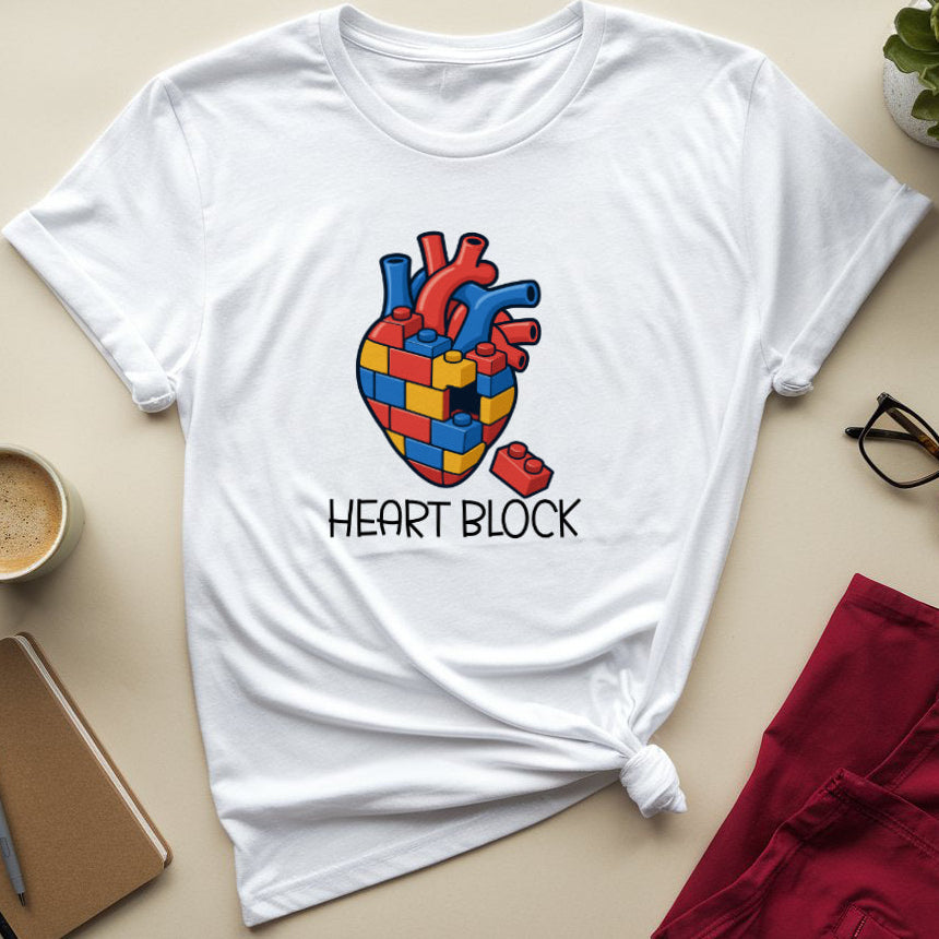 A white short-sleeve graphic T-shirt laid flat on a beige surface, styled with a coffee cup, notebook, pen, glasses, folded red pants, and a small plant. The shirt has a colorful anatomical heart design made of red, blue, and yellow blocks, with the text 'HEART BLOCK' underneath.