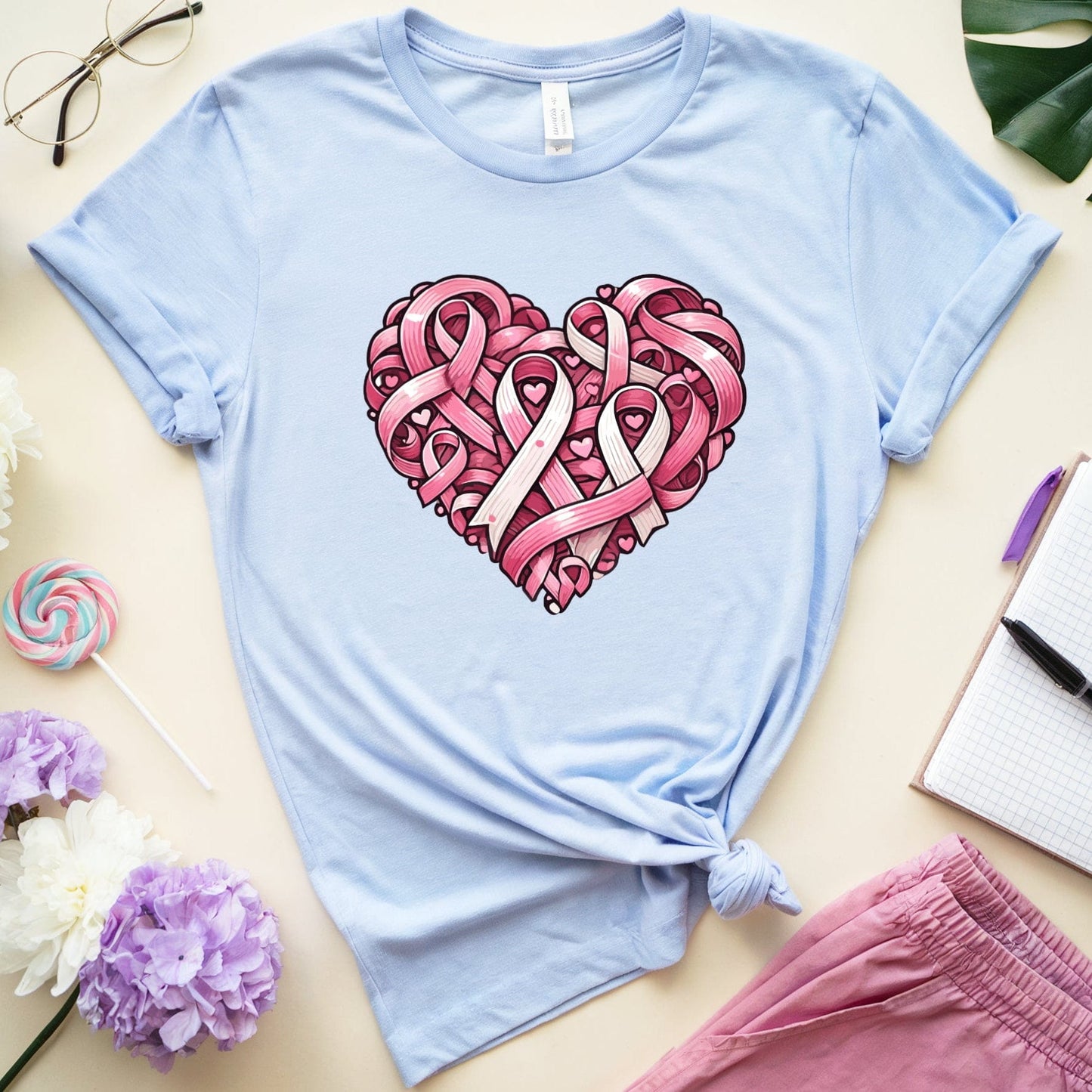 Light blue t-shirt with pink heart design featuring ribbons on a light surface with flowers and glasses.