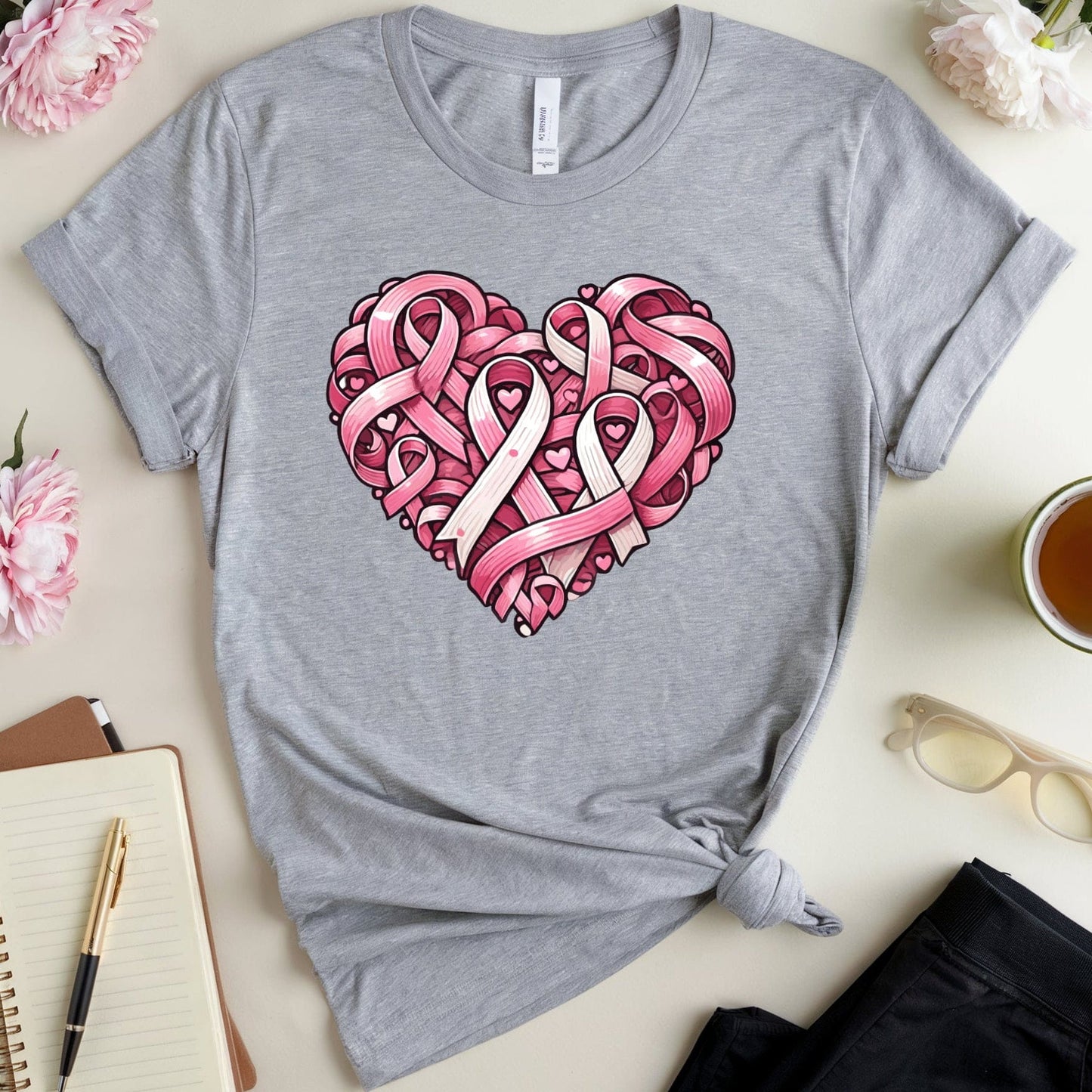 Gray t-shirt with pink heart design featuring ribbons on a light surface with flowers and a cup.
