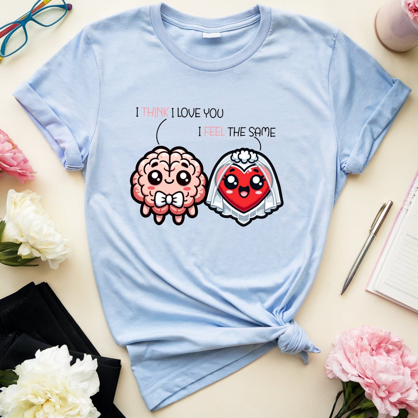 Light blue t-shirt with cartoon brain and heart design on a table with flowers and glasses.