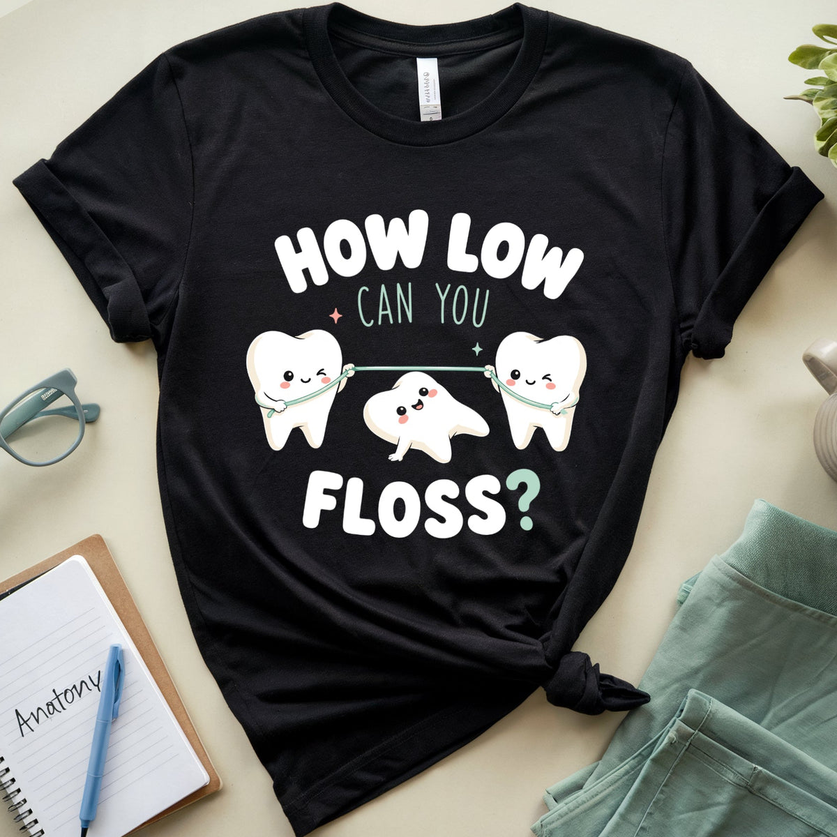A black graphic t-shirt featuring three cartoon teeth holding a floss string, with the text 'HOW LOW CAN YOU FLOSS?' in white and mint green. The shirt is styled in a flat lay on a beige surface with a notebook, glasses, a mug, and mint green shorts.