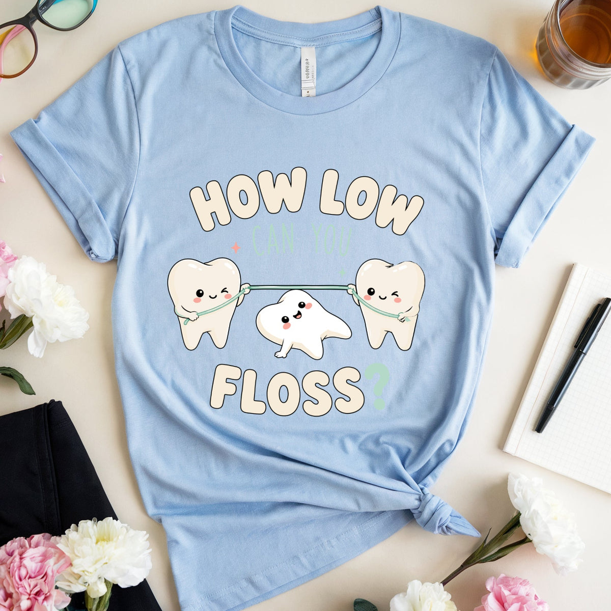 A light blue graphic t-shirt with a cute cartoon design of three teeth pulling a string, with the text 'HOW LOW CAN YOU FLOSS?'. The t-shirt is styled in a flat-lay arrangement with pink and white carnations, eyeglasses, a glass of tea, a notebook, and a pen on a light beige background.