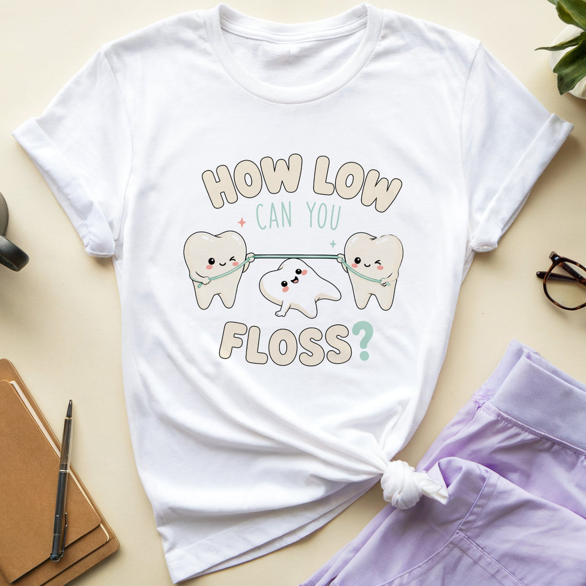 A white graphic t-shirt with a cute cartoon design of three teeth holding hands and the text 'HOW LOW CAN YOU FLOSS?' in beige and mint green. The t-shirt is styled in a flat-lay arrangement on a light beige background, surrounded by a cup of coffee, a notebook, glasses, and lavender pajama pants.