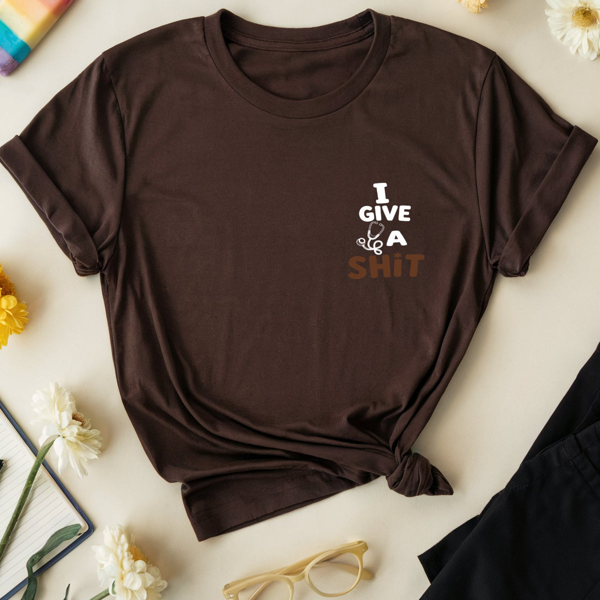 A dark brown short-sleeve t-shirt with a graphic text print reading 'I GIVE U A SHIT' in white and brown, styled in a flat lay with black pants, a rainbow popsicle, yellow and white flowers, glasses, and a notebook on a light beige background.