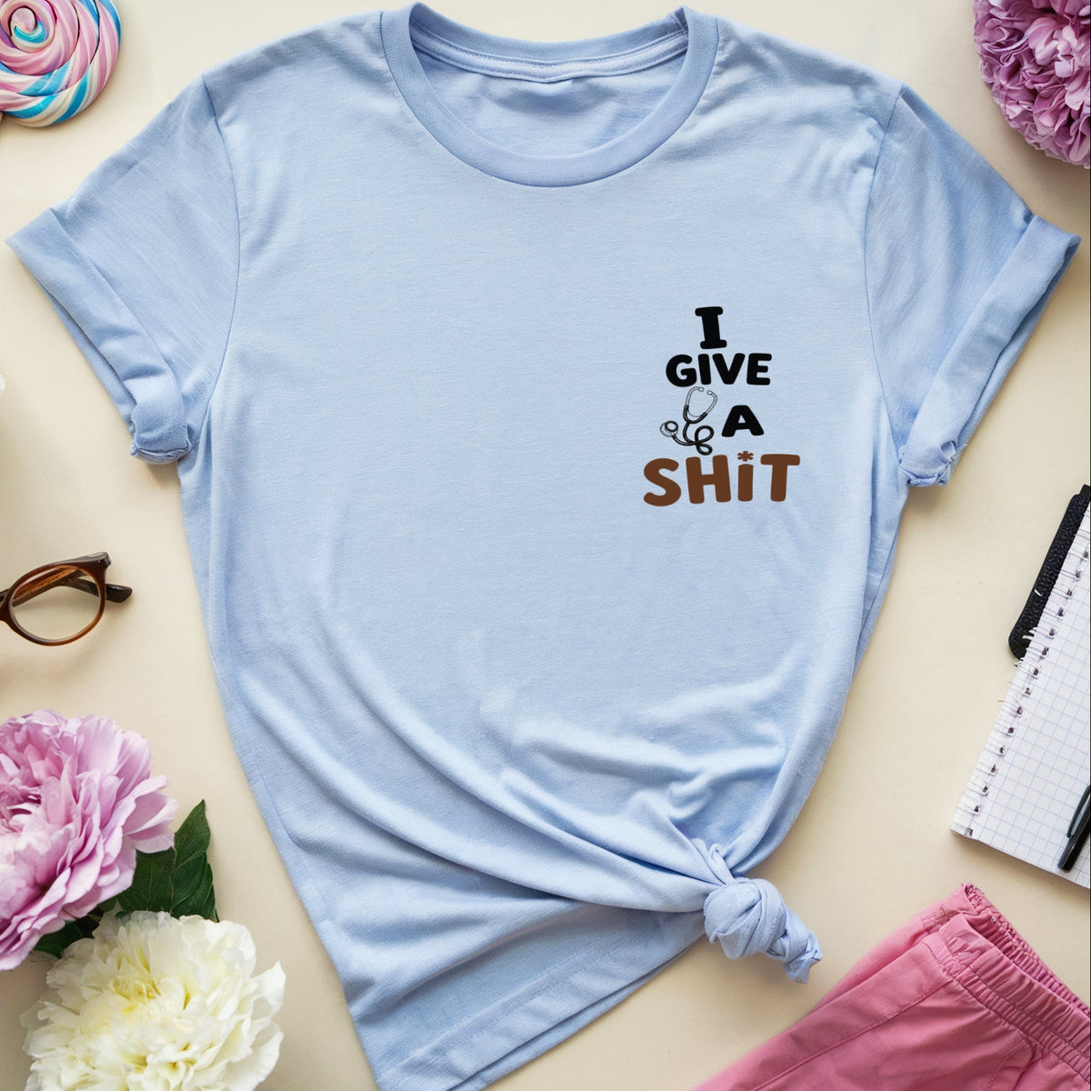 A light blue graphic t-shirt with the text 'I GIVE A SHIT' in black and brown, styled in a flat lay with pink and white peonies, glasses, a notebook, and pink shorts on a beige background.