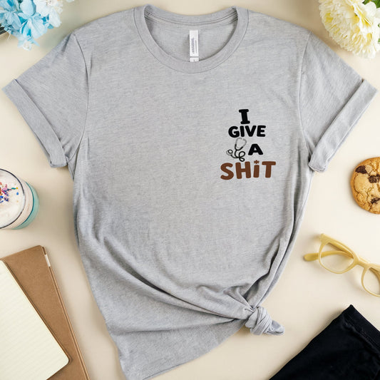 A heather gray short-sleeve t-shirt with the text 'I GIVE A SHIT' printed on the chest, styled in a flat lay with a drink, notebook, flowers, glasses, and a cookie on a light beige background.