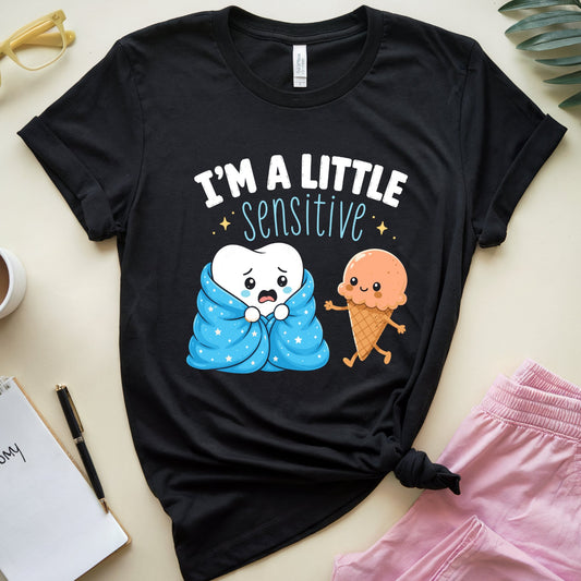 A black graphic t-shirt laid flat on a light beige surface, surrounded by lifestyle props including pink pants, a coffee cup, a notebook, and a plant leaf. The t-shirt features a cartoon graphic of a white tooth wrapped in a blue blanket next to a cute orange ice cream cone, with the text 'I'M A LITTLE sensitive'.