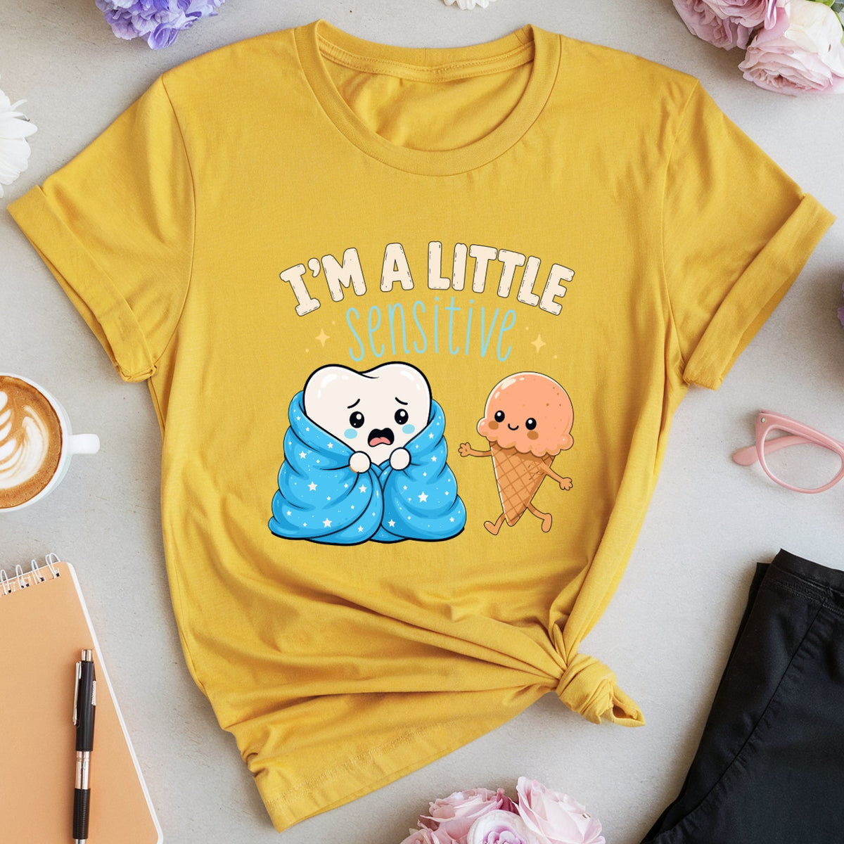 A yellow graphic t-shirt with a cartoon design of a sad tooth and an ice cream cone, surrounded by lifestyle flat-lay props including flowers, a notebook, glasses, and folded black pants on a light gray background.