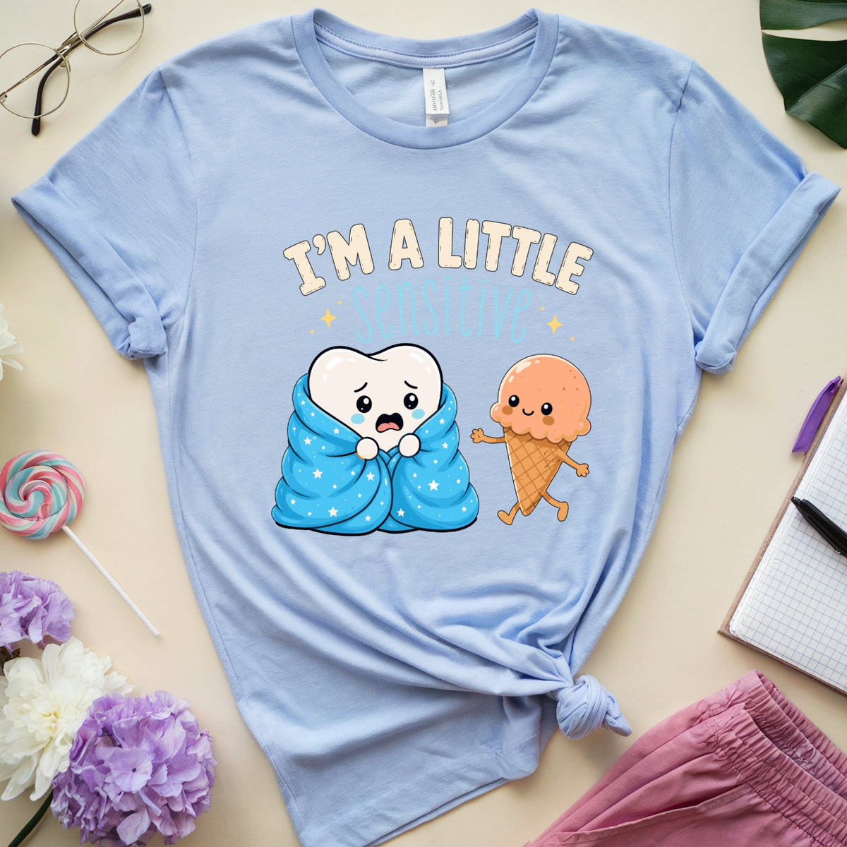 A light blue graphic t-shirt laid flat on a beige surface, styled with flowers, glasses, a notebook, and pink pants. The shirt features a cartoon graphic of a tooth wrapped in a blue blanket and an ice cream cone, with the text 'I'M A LITTLE SLEEPER'.