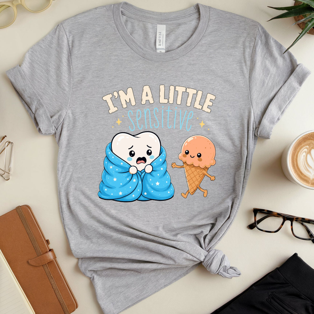 A heather gray graphic t-shirt with a cute cartoon design of a tooth wrapped in a blue blanket and an ice cream cone character. The text reads 'I'M A LITTLE SPISSED!' in beige and light blue.