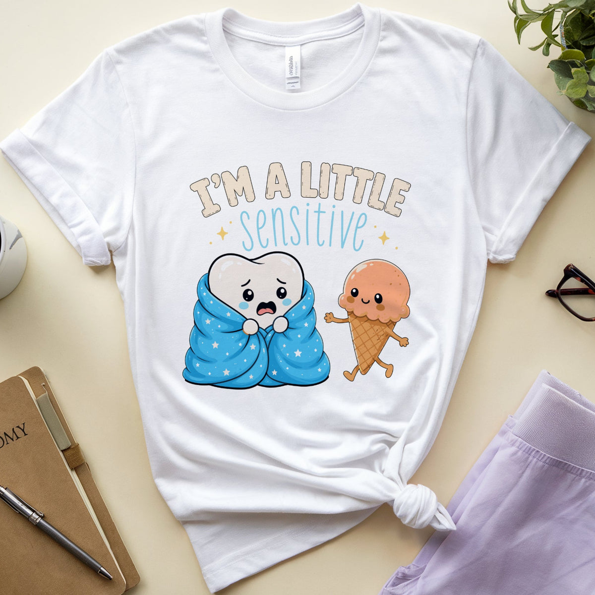 A white graphic t-shirt with a cute cartoon design of a baby tooth wrapped in a blue blanket next to an ice cream cone, with the text 'I'M A LITTLE SENSITIVE'. The t-shirt is styled in a flat-lay arrangement on a beige background with a notebook, glasses, a cup of coffee, and a plant.