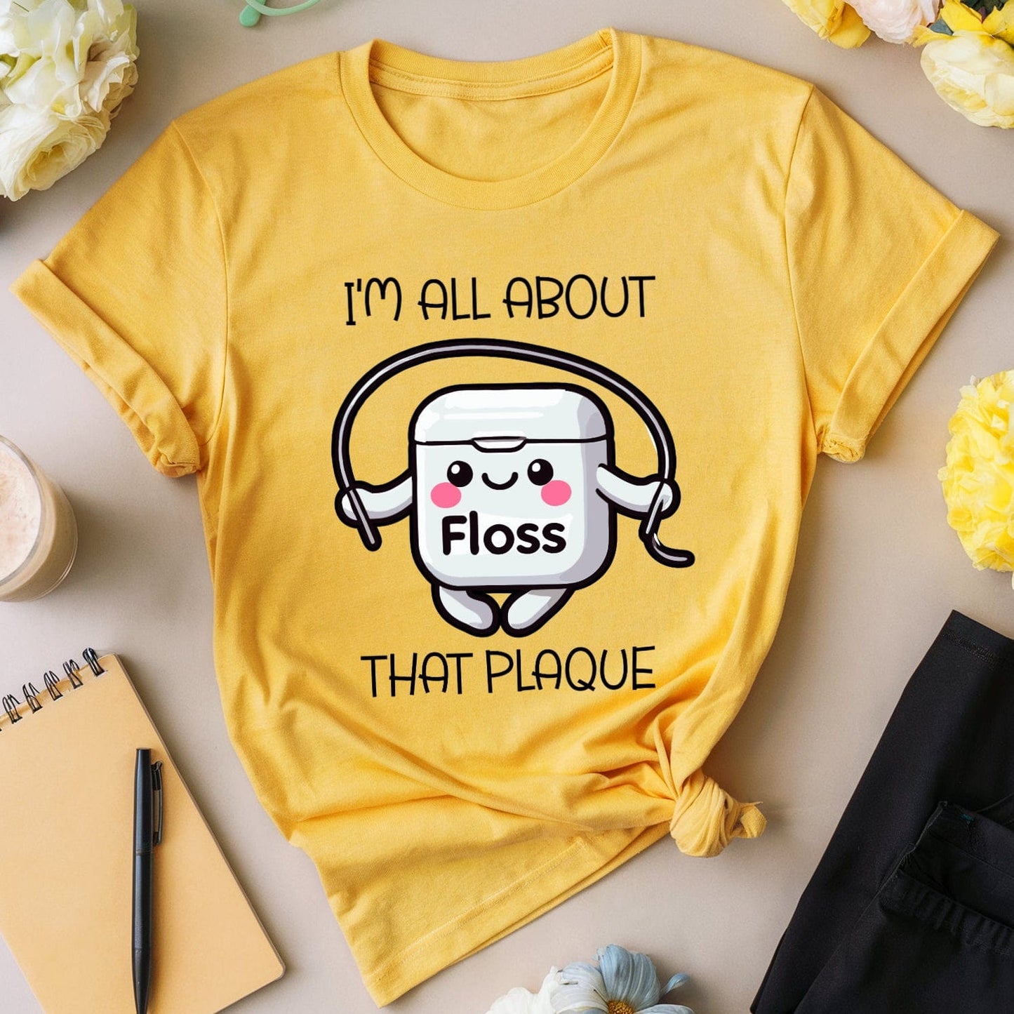 Yellow t-shirt with dental floss graphic and text, surrounded by flowers and stationery on a light surface.