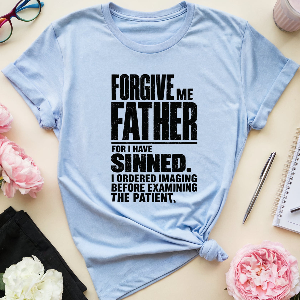 A light blue graphic t-shirt with rolled sleeves and a tied hem, featuring black text that reads 'FORGIVE ME FATHER FOR I HAVE SINNED. I ORDERED IMAGING BEFORE EXAMINING THE PATIENT.
