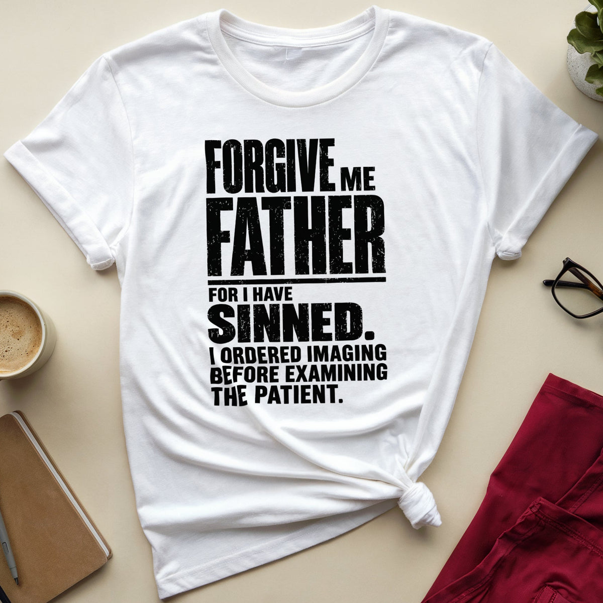A white short-sleeve graphic T-shirt laid flat on a beige surface, surrounded by lifestyle props including a coffee cup, notebook, glasses, folded red pants, and a small plant. The shirt has large black text reading, 'FORGIVE ME FATHER FOR I HAVE SINNED.