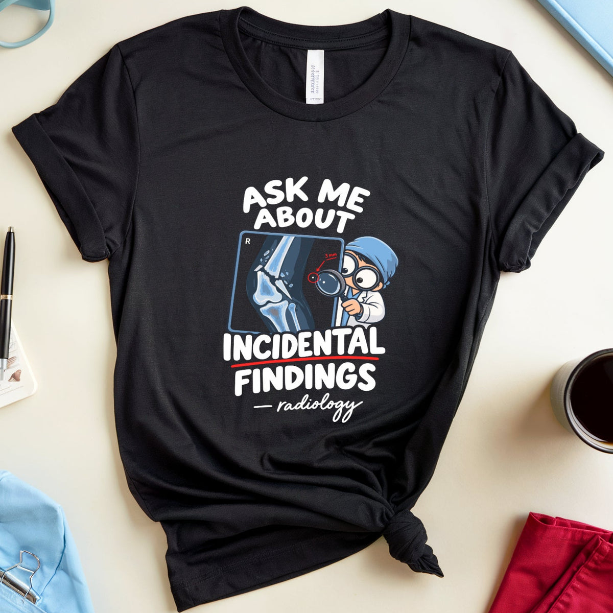 A black graphic t-shirt with a medical/nursing theme, styled in a flat-lay arrangement on a light beige background with blue glasses, a anatomy book, a cup of coffee, and medical scrubs.