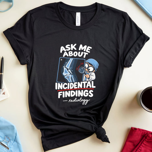 A black graphic t-shirt with a medical/nursing theme, styled in a flat-lay arrangement on a light beige background with blue glasses, a anatomy book, a cup of coffee, and medical scrubs.