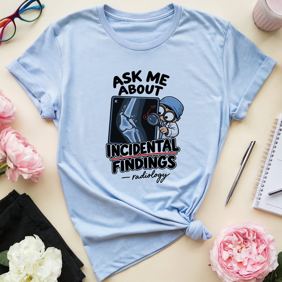 A light blue graphic t-shirt with a tie knot, featuring a radiology-themed print that reads 'ASK ME ABOUT INCIDENTAL FINDINGS - radiology'. The t-shirt is styled in a flat-lay arrangement on a beige background with pink flowers, glasses, a drink, a notebook, and a pen.