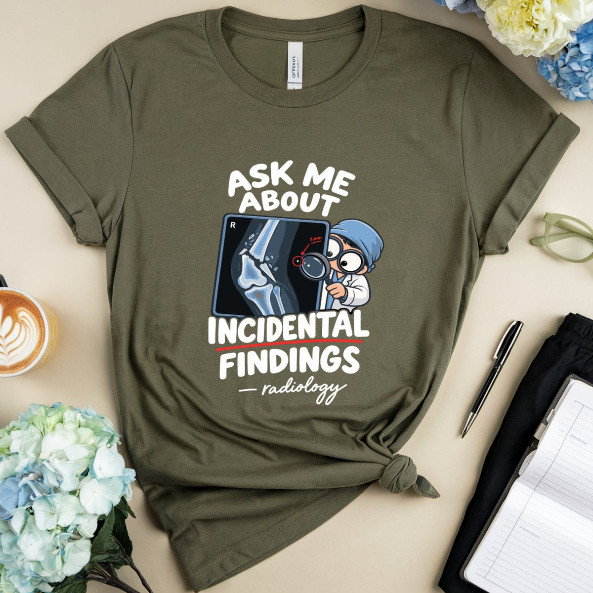 An olive green graphic t-shirt is presented in a styled flat-lay arrangement on a beige background. The shirt features a white text graphic reading 'ASK ME ABOUT INCIDENTAL FINDINGS - radiology' alongside a cartoon illustration of a radiologist.