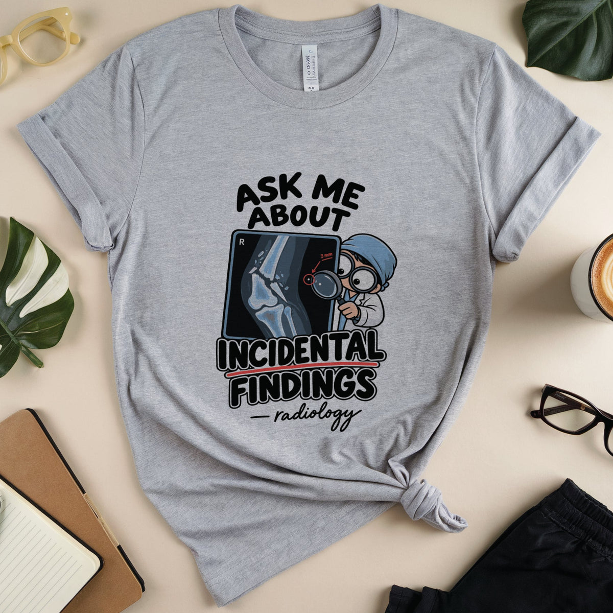 A heather gray graphic t-shirt with a radiology-themed print reading 'ASK ME ABOUT INCIDENTAL FINDINGS - radiology', styled in a flat lay with glasses, a coffee cup, notebook, and plant leaves on a beige background.