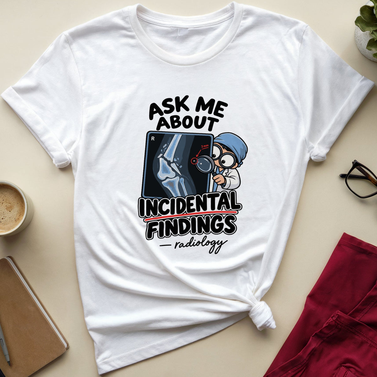 A white graphic t-shirt with a radiology-themed print reading 'ASK ME ABOUT INCIDENTAL FINDINGS - radiology' is styled in a flat lay on a beige surface with a coffee cup, notebook, glasses, red pants, and a small plant.
