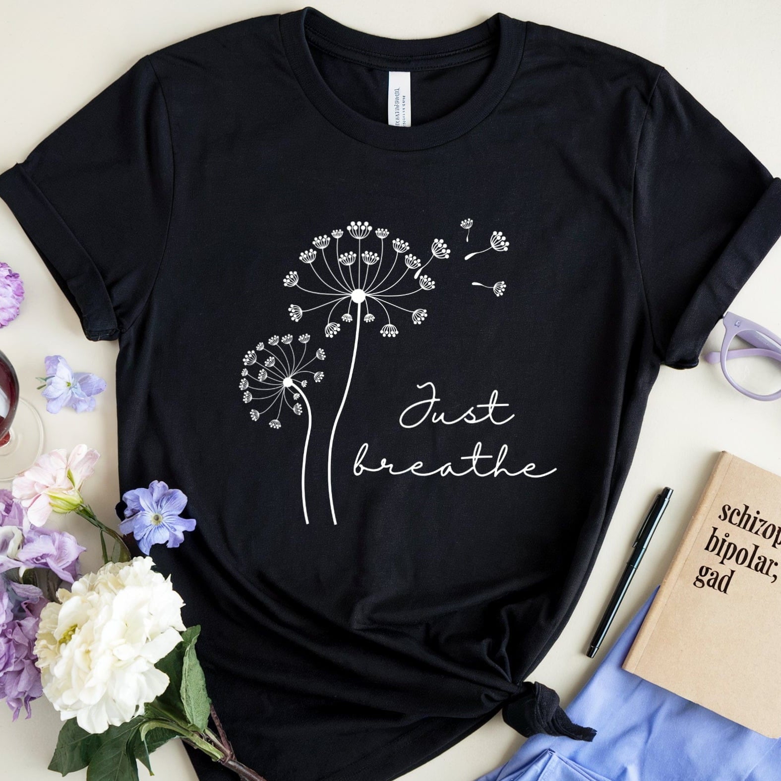 Black t-shirt with dandelion design and 'Just breathe' text on a white surface with flowers and glasses.