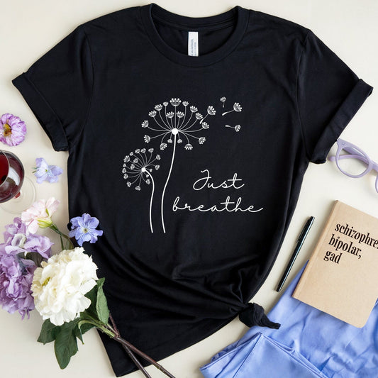 Black t-shirt with dandelion design and 'Just breathe' text on a white surface with flowers and glasses.