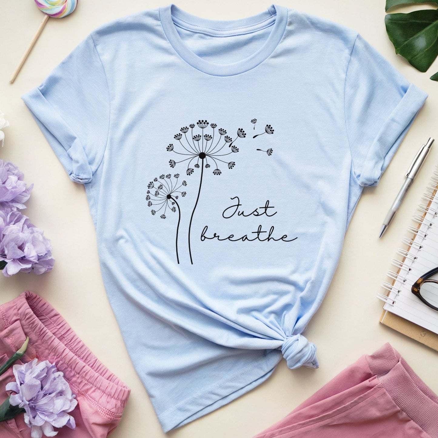 Light blue t-shirt with dandelion graphic and 'Just breathe' text on a light surface with flowers and accessories.