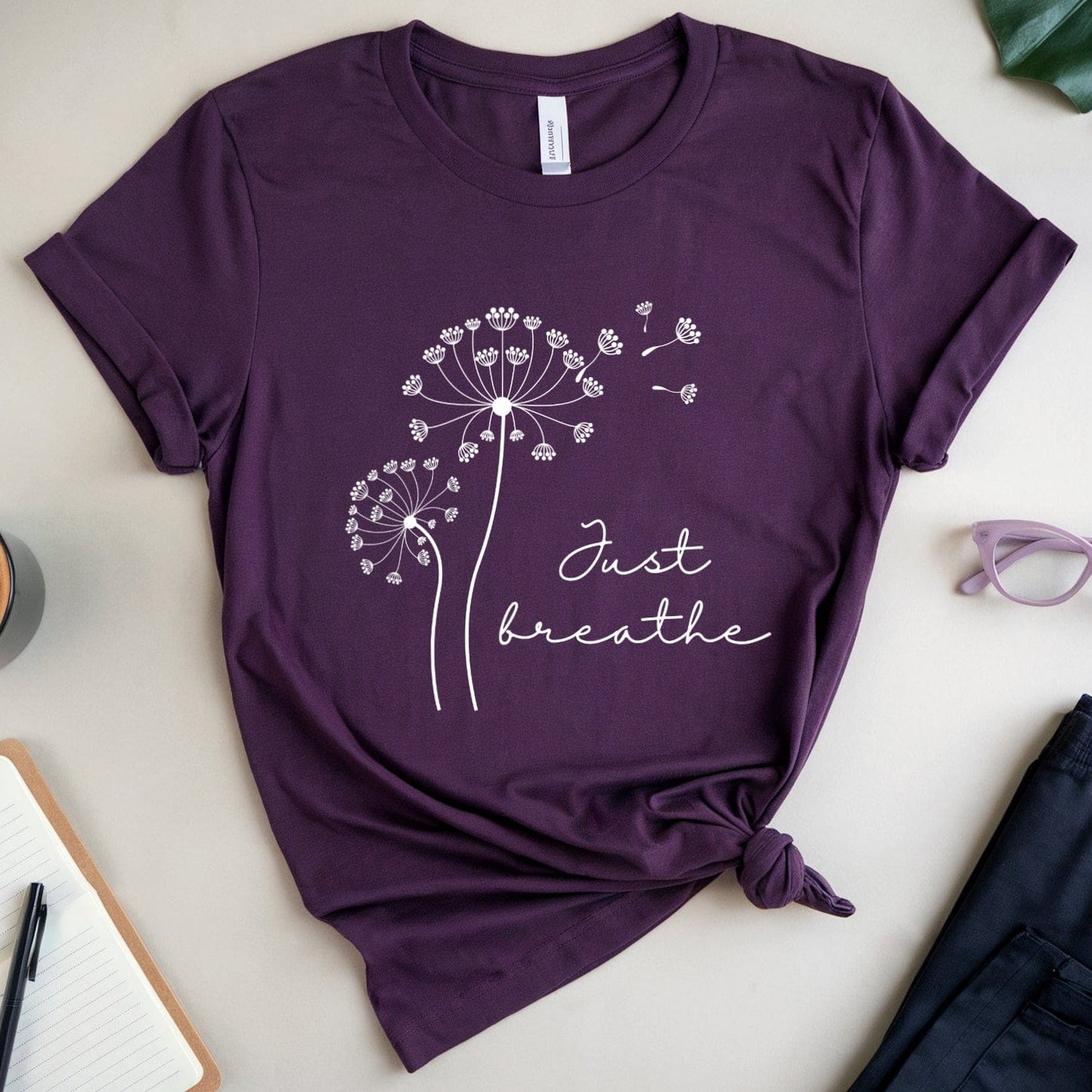 Purple t-shirt with dandelion design and 'Just breathe' text on a light surface with coffee, glasses, and notebook.