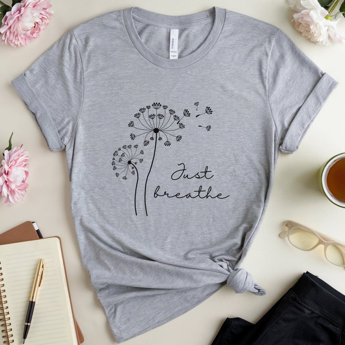 Gray t-shirt with dandelion design and 'Just breathe' text on a table with flowers and a notebook.
