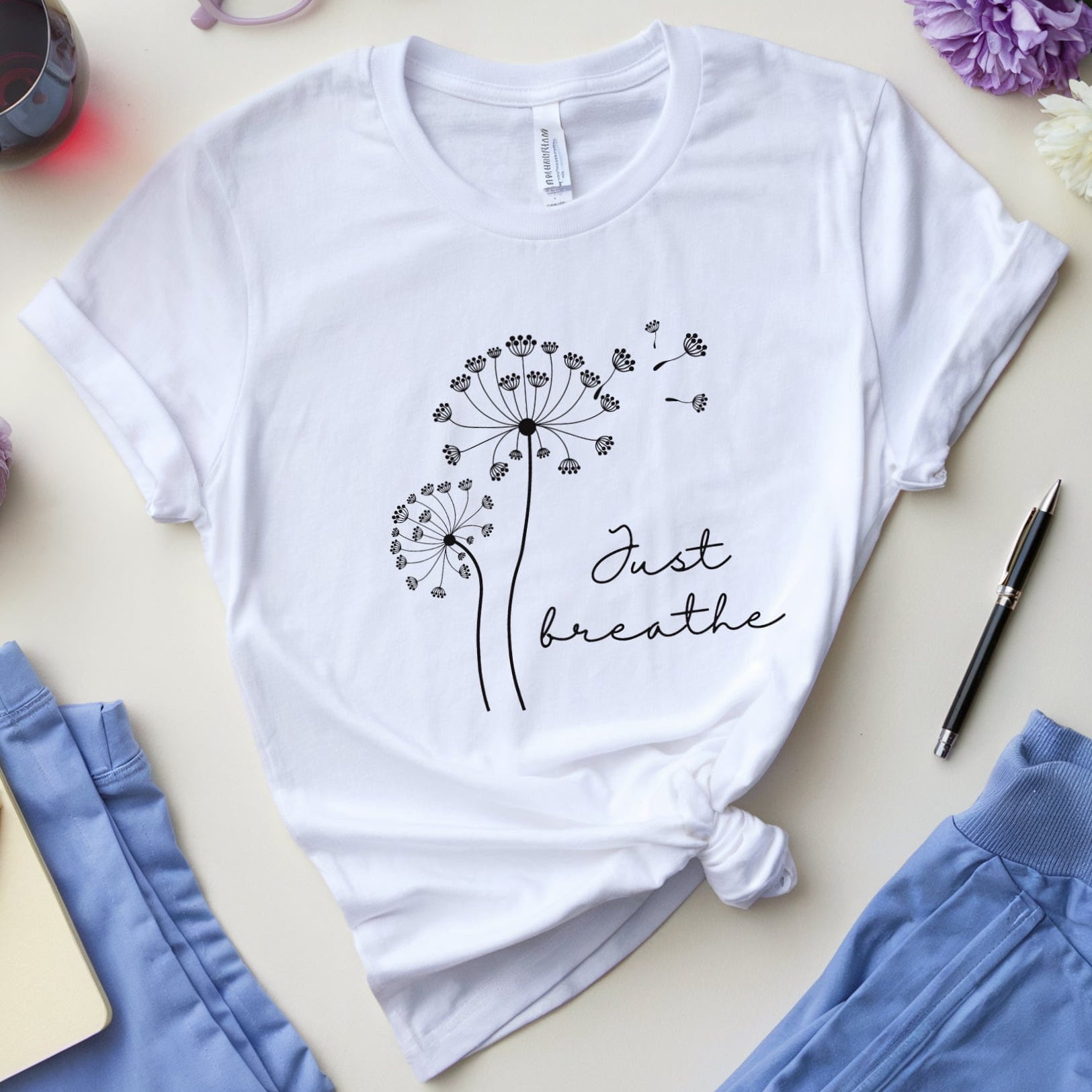 White t-shirt with dandelion design and 'Just breathe' text on a table with flowers and glasses.